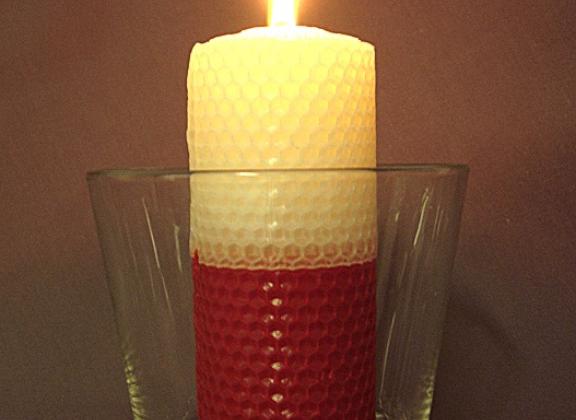 Kuvassa punavalkoinen mehiläisvahakynttilä lasimaljassa. Kynttilässä on tuli.